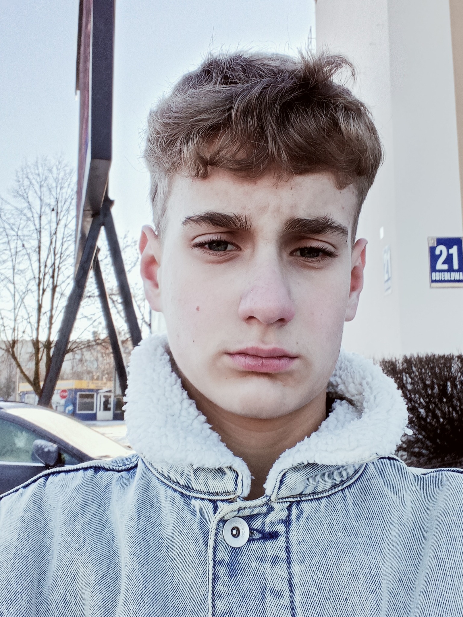 Młody mężczyzna w dżinsowej kurtce z futrzanym kołnierzem, na tle budynku z numerem 21 i drzew bez liści.