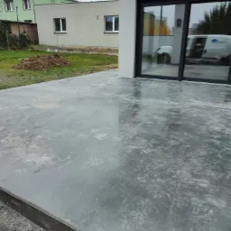 Świeżo wylana, szara, betonowa wylewka tarasu z widocznymi liniami dylatacyjnymi, tuż przy nowoczesnym domu z dużymi przesuwnymi drzwiami tarasowymi, w których odbija się biały samochód dostawczy.