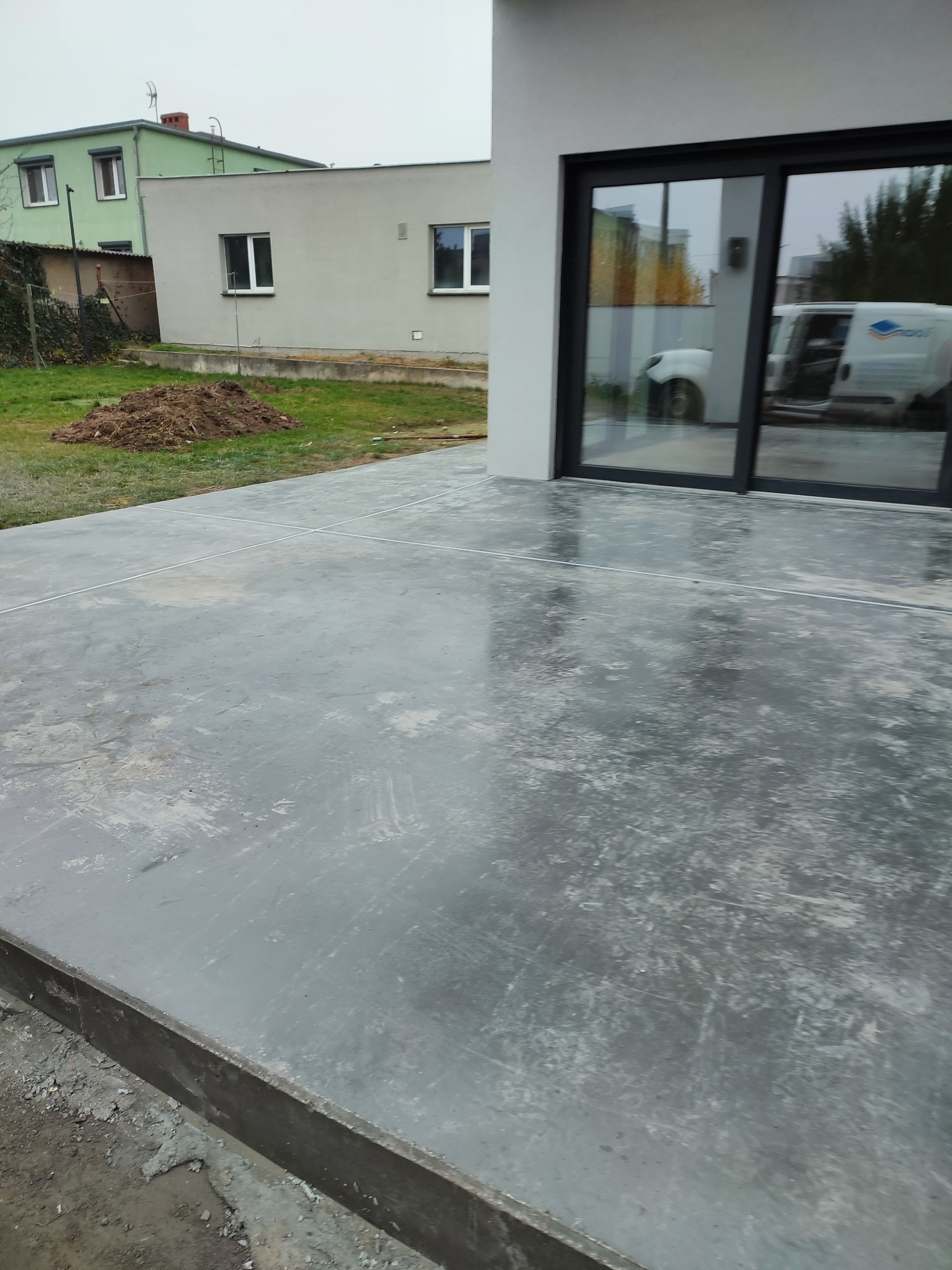 Świeżo wylana, szara, betonowa wylewka tarasu z widocznymi liniami dylatacyjnymi, tuż przy nowoczesnym domu z dużymi przesuwnymi drzwiami tarasowymi, w których odbija się biały samochód dostawczy.