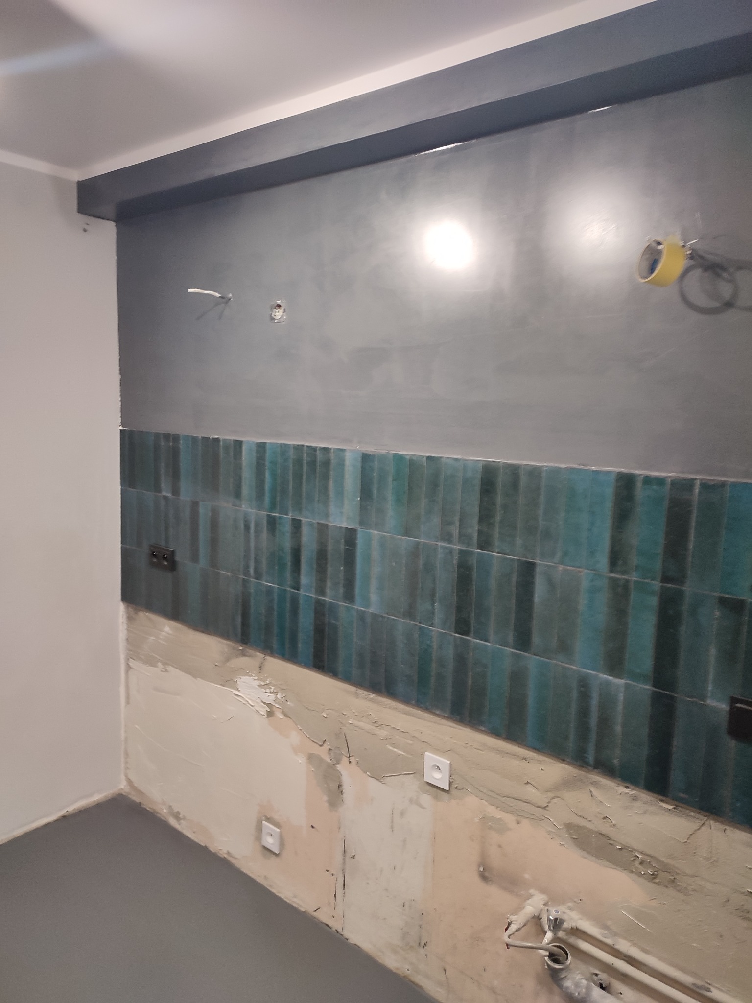 Ściana w trakcie remontu z położonym pasem pionowych, turkusowych płytek, widoczne wystające kable, gładź i szpachla, szara farba na górnej części ściany i suficie.