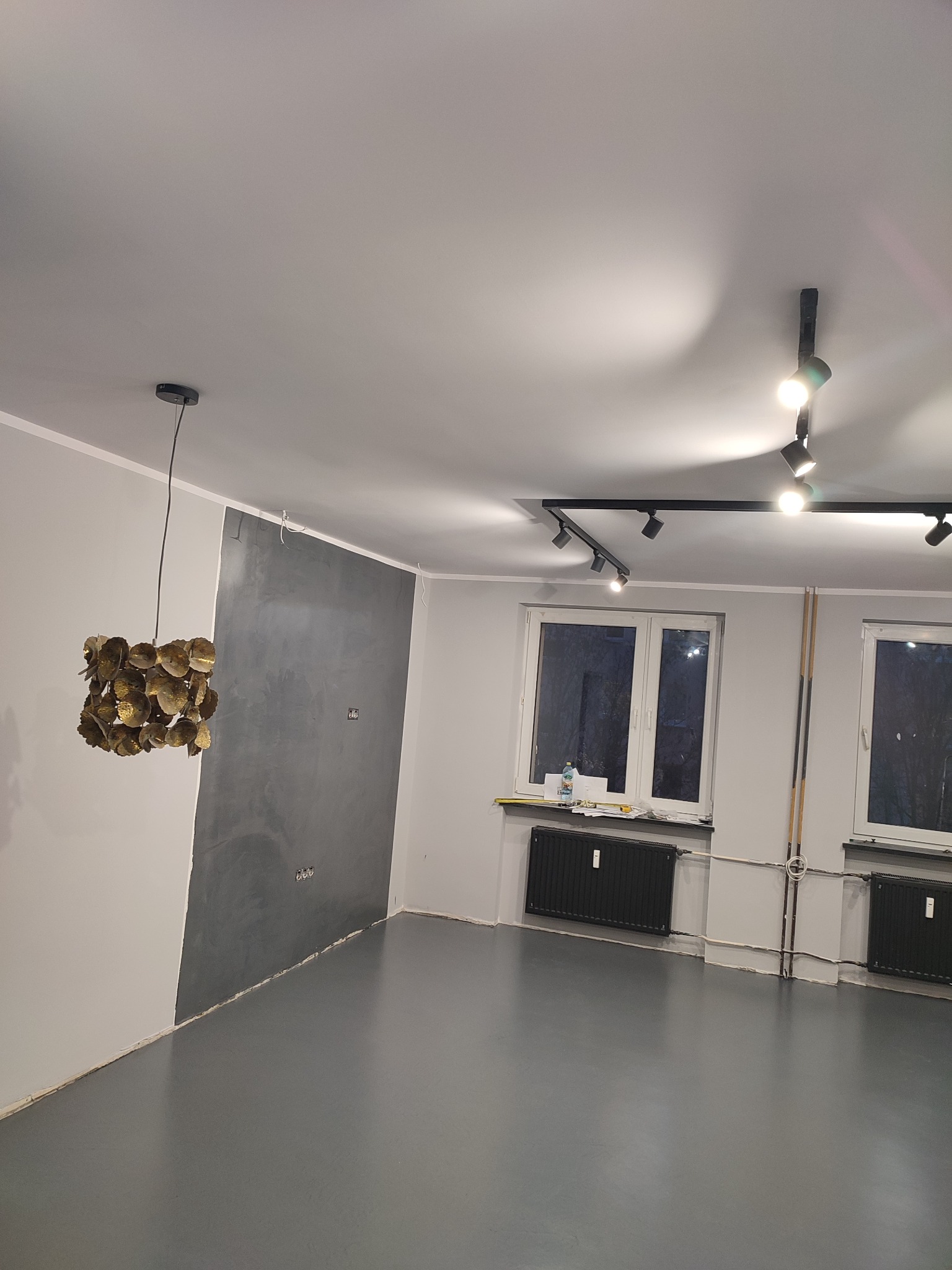 Półmatowa szara podłoga w pustym pokoju po remoncie, z fragmentem ściany pokrytym dekoracyjną szarą farbą, nowoczesnym oświetleniem szynowym i złotą ozdobną lampą wiszącą.