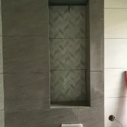 Ściana z szarych i białych płytek z wnęką wyłożoną płytkami w wzór liści, poziomica oparta o ścianę, widoczne niedokończone gniazdko elektryczne.