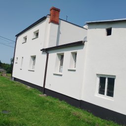 GOLDEN-BUD - Biała elewacja domu z ceglanym kominem i brązowymi rynnami, widok z poziomu trawnika w słoneczny dzień. Teksturowana powierzchnia ścian.