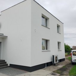 GOLDEN-BUD - Dwukondygnacyjny dom po renowacji elewacji, pomalowany na biało z ciemnoszarą podmurówką. Widoczny fragment dachu, okna i firmowy bus z reklamą usług.