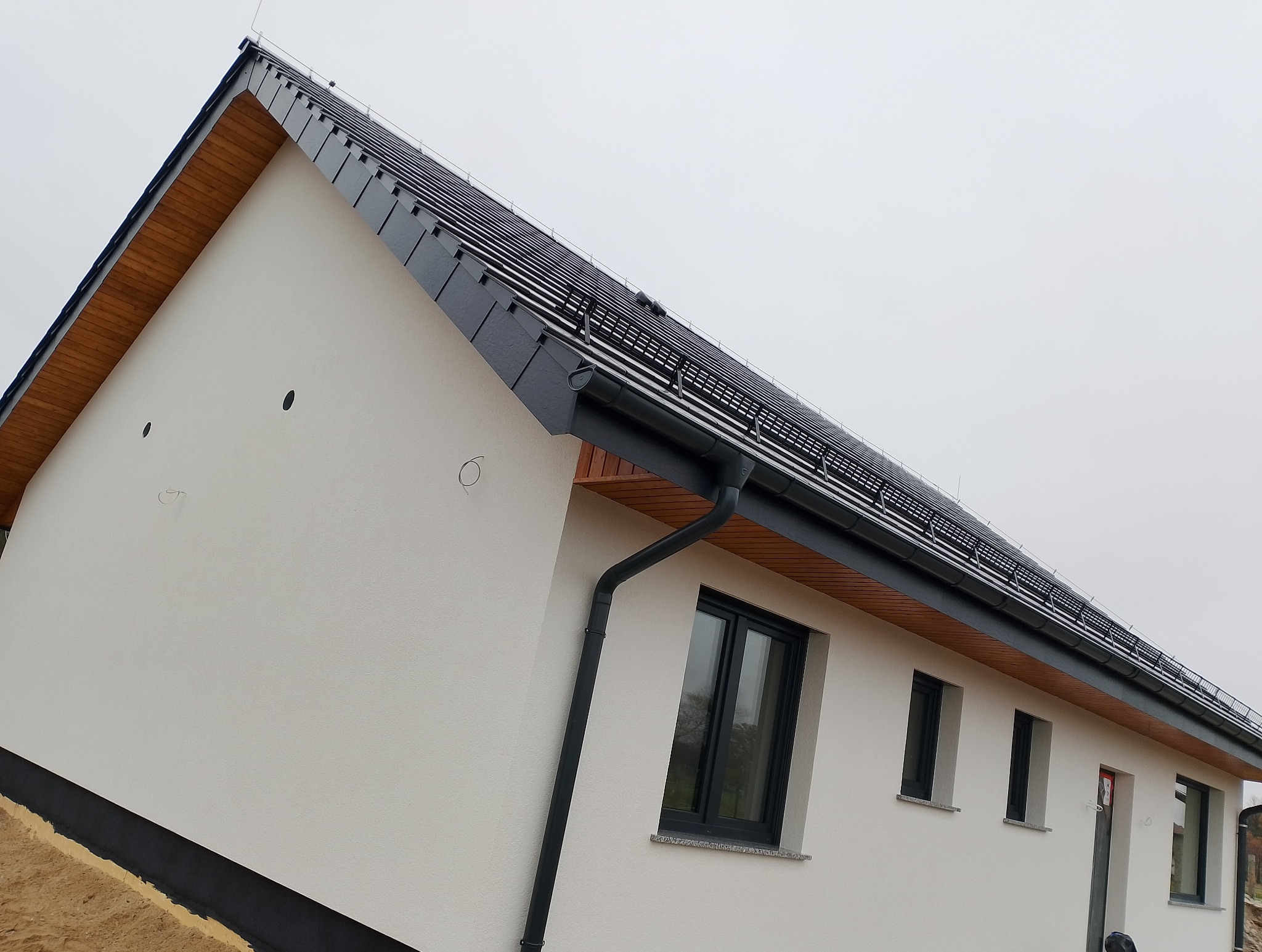 Nowoczesny dom jednorodzinny z jasną elewacją, czarną dachówką i orynnowaniem. Widoczne okna i fragment dachu. Ujęcie z dołu podkreśla geometrię budynku.