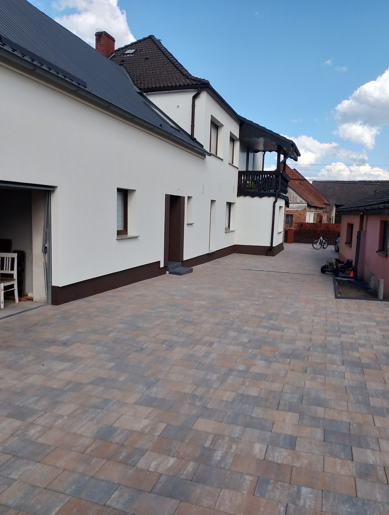 Biały dom z ciemnym dachem i balkonem, widok z poziomu kostki brukowej w odcieniach szarości i brązu. Błękitne niebo z chmurami w tle.