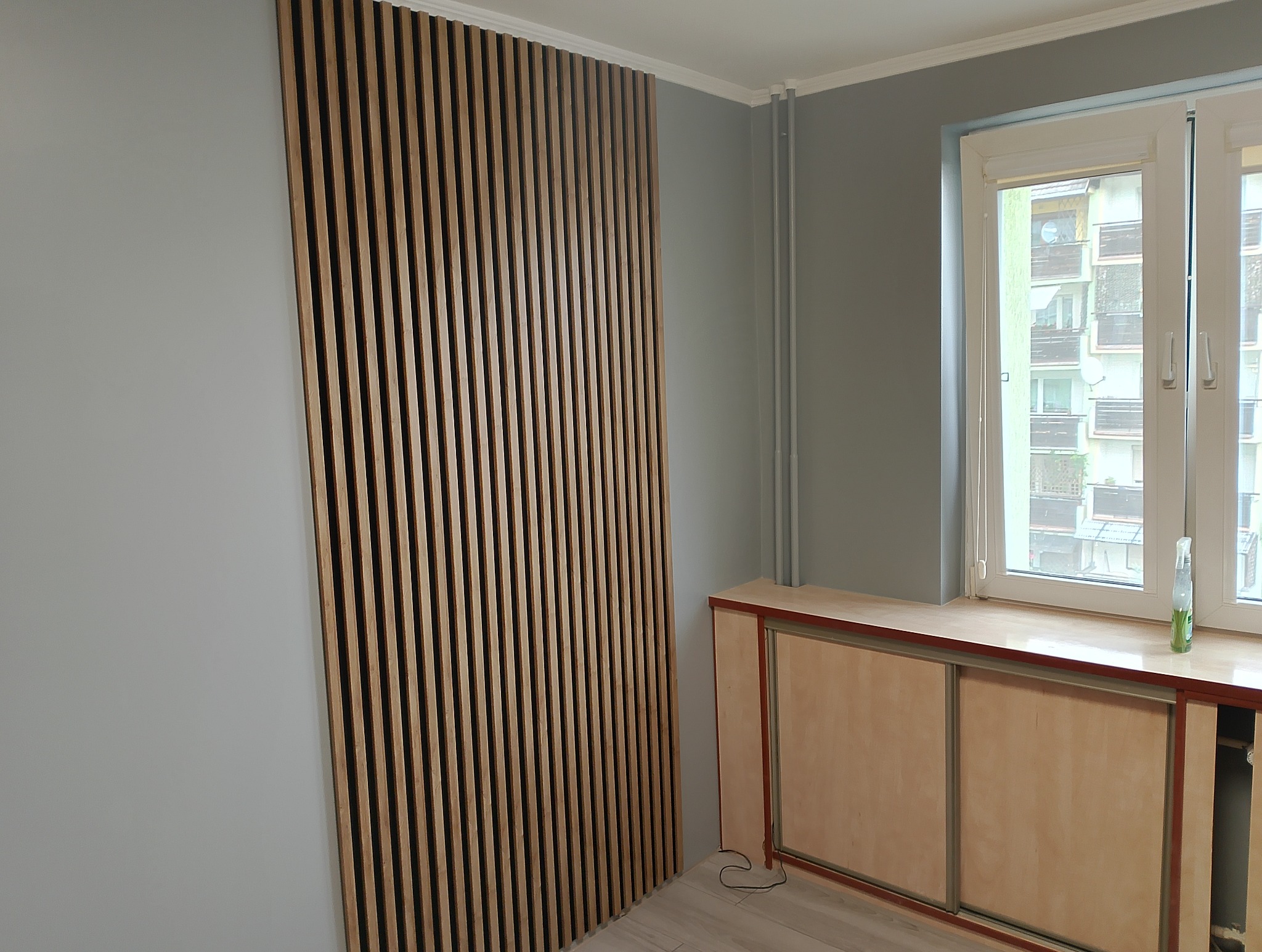 Drewniane lamele na ścianie w szarym pokoju, widok na okno z zabudową. Nowoczesny design wnętrza, pionowe linie, jasne drewno, minimalistyczny styl.