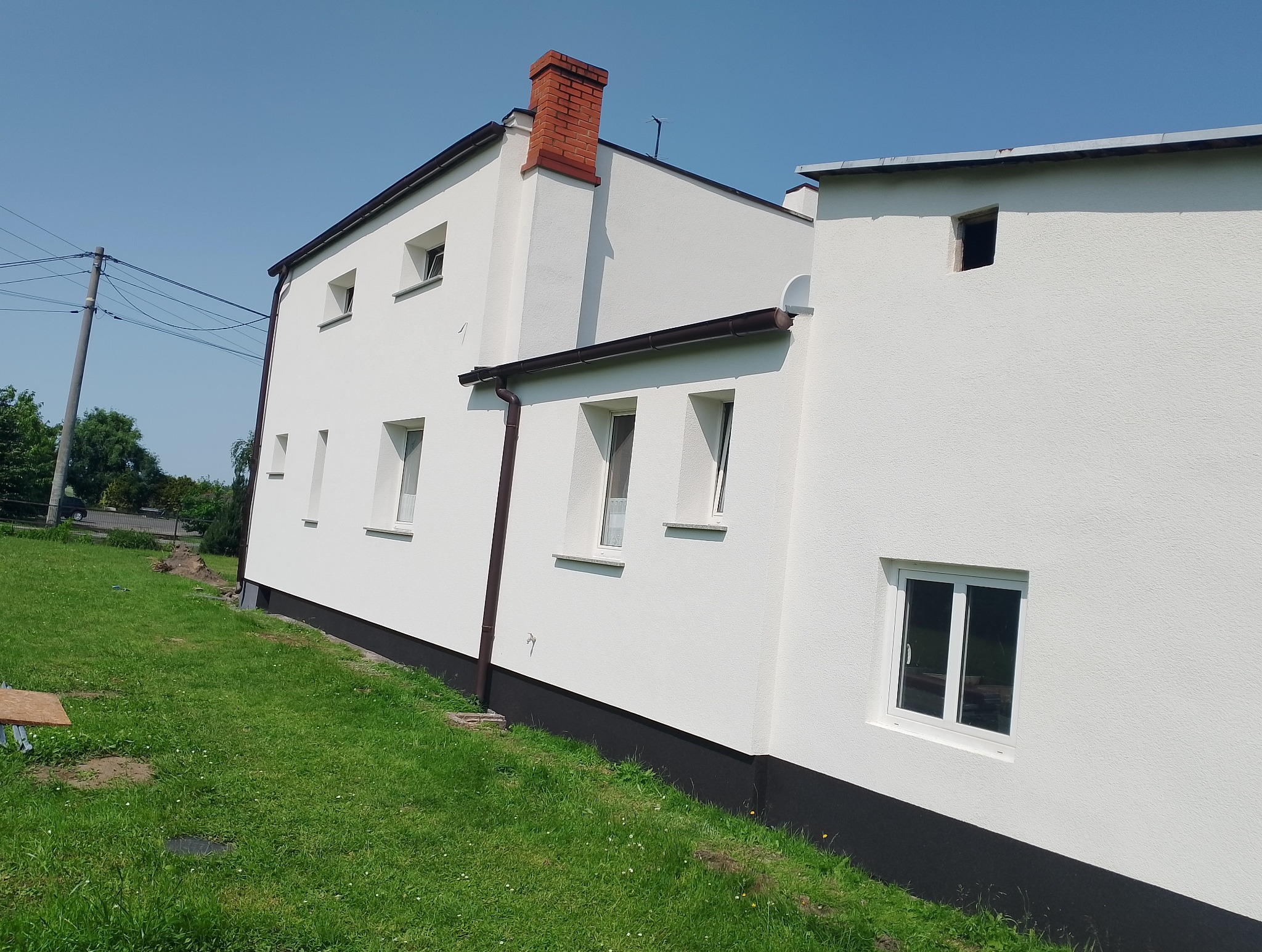 Biała elewacja domu z ceglanym kominem i brązowymi rynnami, widok z poziomu trawnika w słoneczny dzień. Teksturowana powierzchnia ścian.