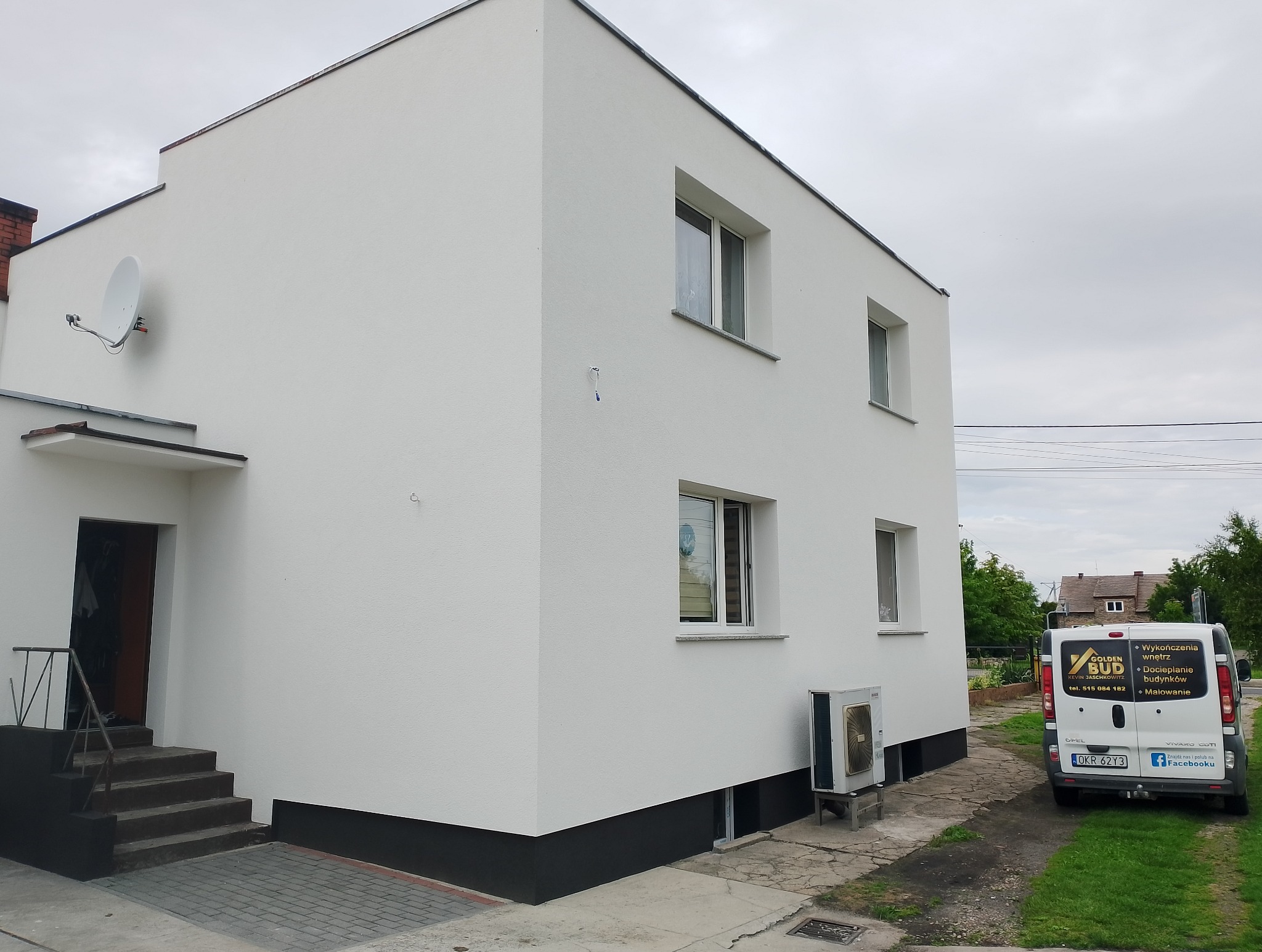 Dwukondygnacyjny dom po renowacji elewacji, pomalowany na biało z ciemnoszarą podmurówką. Widoczny fragment dachu, okna i firmowy bus z reklamą usług.