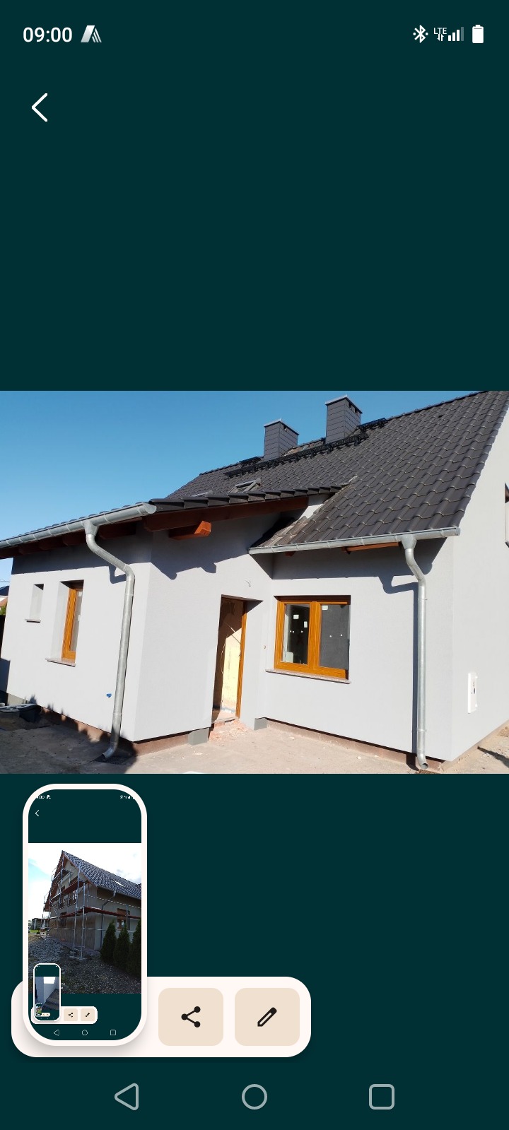 Otynkowany dom jednorodzinny z brązowymi oknami i szarą dachówką, widoczny z zewnątrz w słoneczny dzień. Na ekranie telefonu widoczne rusztowania na wcześniejszym etapie.