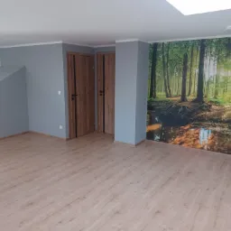 Pusty pok&oacute;j po remoncie z szarą ścianą, dwojgiem drewnianych drzwi z czarnymi paskami, panele podłogowe i fototapeta z motywem leśnym.