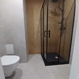 Nowoczesna łazienka z kabiną prysznicową w czarnej ramie, wiszącą toaletą i brązowymi płytkami z geometrycznym wzorem na ścianie.