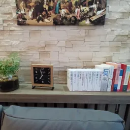 Gabinet terapeutyczny z książkami o tematyce psychologicznej, zegarem w drewnianej ramce i rośliną doniczkową na tle ściany z kamienia dekoracyjnego, nad którą wisi reprodukcja obrazu.