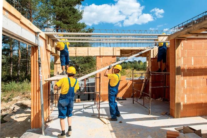 Budowa domu z czerwonej cegły: murarze w żółtych koszulkach i niebieskich kombinezonach montują metalowe belki stropowe na tle błękitnego nieba z chmurami.
