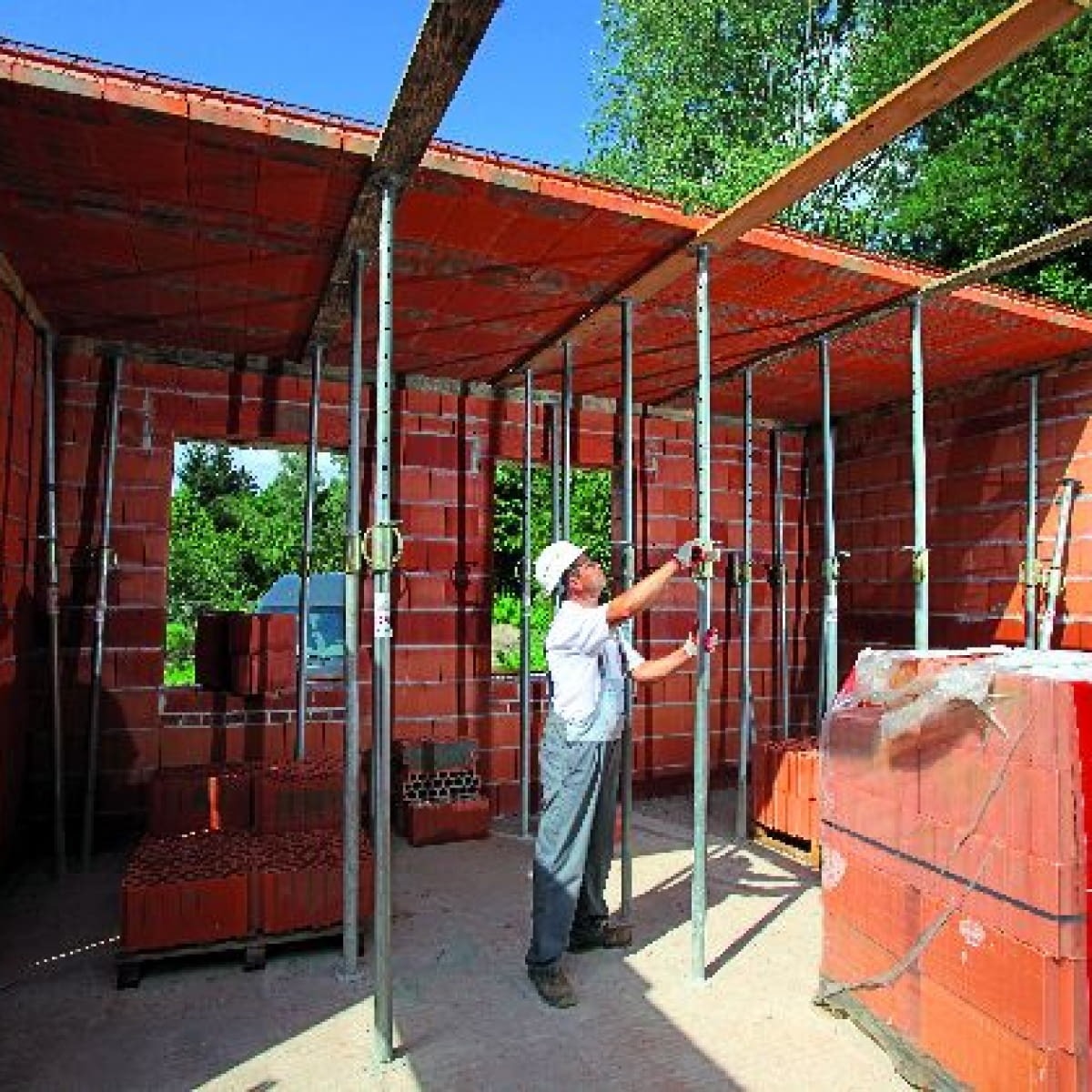 Budowa domu z czerwonej cegły: murarz w kasku ustawia podpory stropu, w tle widoczne pakiety cegieł i drzewa za oknem.
