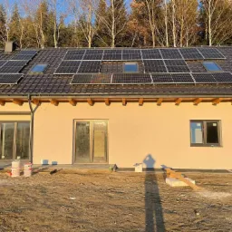 Nowoczesny dom jednorodzinny z panelami słonecznymi na ciemnym dachu, widoczne okna dachowe, świeżo otynkowana elewacja w odcieniach beżu, teren wokół domu w trakcie prac wykończeniowych, widoczne...