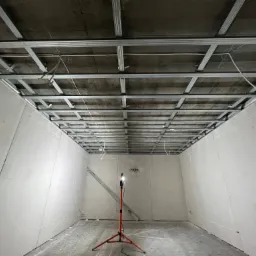 Montaż metalowej konstrukcji sufitu podwieszanego z płyt karton-gips, widoczne kable elektryczne, ściany wykończone płytami gipsowymi, lampa robocza na trójnogu i poziomica oparta o ścianę.