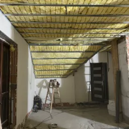 Montaż izolacji z wełny mineralnej na metalowym stelażu sufitu podwieszanego, widoczna drabina i narzędzia w surowym wnętrzu w trakcie prac wykończeniowych.