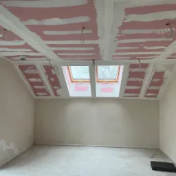 Poddasze w trakcie prac wykończeniowych: widoczne płyty w kolorze różowym na suficie i ścianach, częściowo pokryte białą masą szpachlową, dwa okna dachowe i wystające kable elektryczne.