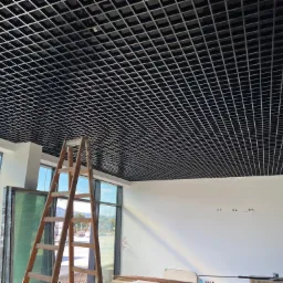 Montaż czarnego sufitu rastrowego w jasnym pomieszczeniu z dużymi oknami, widoczna drewniana drabina, przygotowane okna do montażu i wystające kable elektryczne ze ściany.