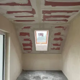 Poddasze w trakcie prac wykończeniowych: widoczne szpachlowane łączenia płyt na suficie i ścianach, okno dachowe i surowa podłoga z zabrudzeniami po pracach budowlanych. Na oknie naklejone ulotki.