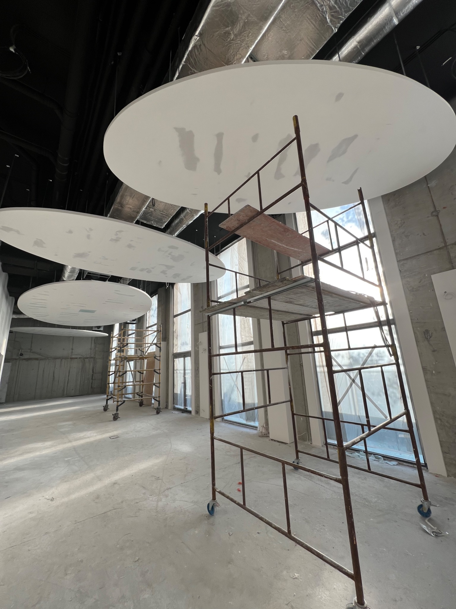 Montaż okrągłych podwieszanych elementów sufitowych w surowym wnętrzu biurowym, widoczne rusztowania, duże okna z widokiem na zewnątrz, instalacja pod sufitem