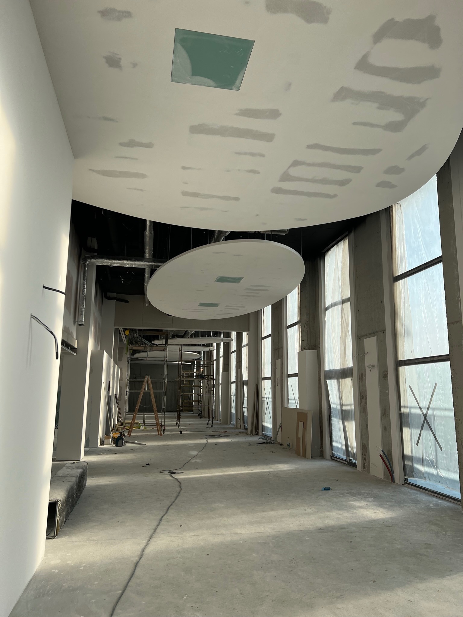 Wnętrze budynku w trakcie prac wykończeniowych: widoczne surowe ściany, okna zabezpieczone folią, fragmenty instalacji, metalowe rusztowania oraz podwieszane, okrągłe elementy sufitu z widocznymi...