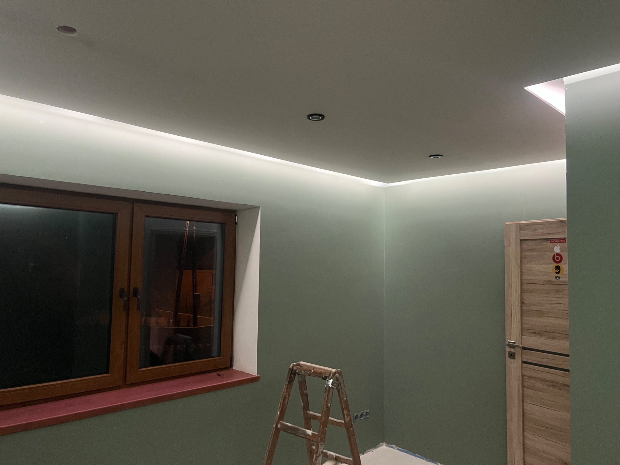 Wnętrze pokoju w trakcie remontu, świeżo pomalowane ściany w odcieniach zieleni, drewniane okno z ciemną ramą i parapetem, drabina malarska, sufit z oświetleniem LED wzdłuż krawędzi i punktowymi...