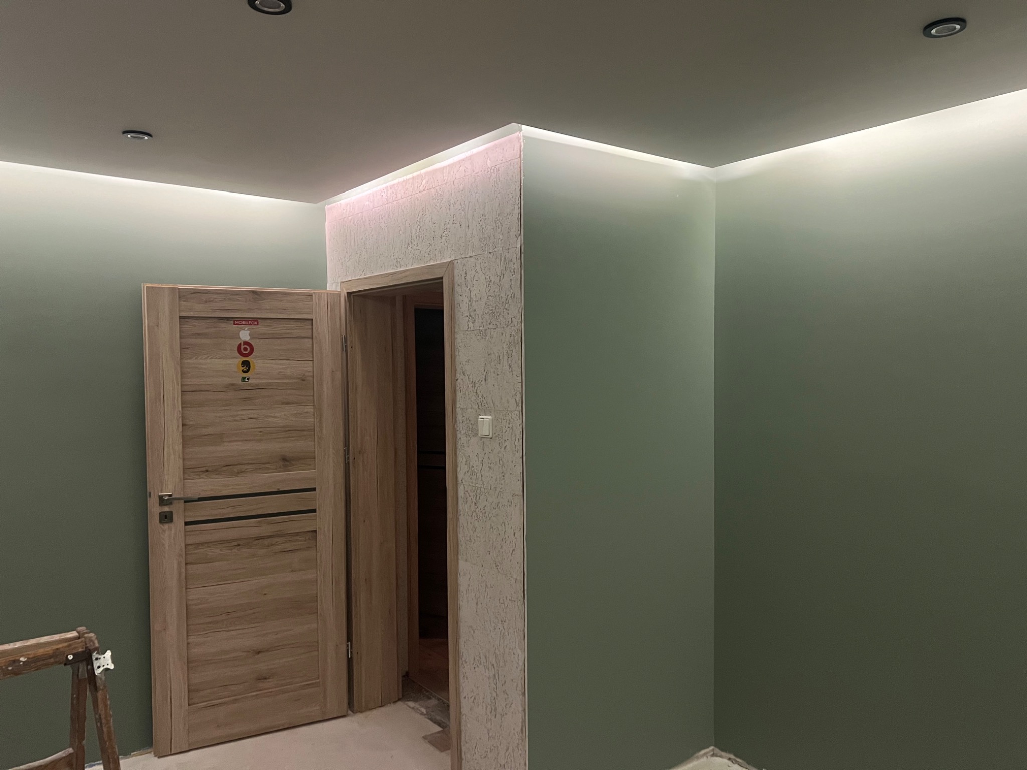 Pomieszczenie po remoncie z oświetleniem LED podsufitowym, pomalowanymi na oliwkowo ścianami, drewnianymi drzwiami z naklejkami i wykończonym wejściem do drugiego pokoju.