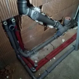 Fragment ściany z cegły dziurawki, przez kt&oacute;rą przechodzą rury instalacji wodnej i kanalizacyjnej, częściowo zaizolowane czerwoną pianką, z widocznymi elementami łączeniowymi i uszczelnieniami.