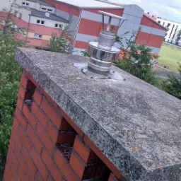 Ceglany komin z betonową czapą i metalowym wkładem kominowym, widok z góry na tle budynków i zieleni.