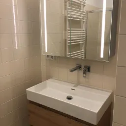 Nowoczesna łazienka z białą umywalką nablatową na drewnianej szafce, chromowaną armaturą i lustrem z oświetleniem LED. W tle widoczny grzejnik drabinkowy.