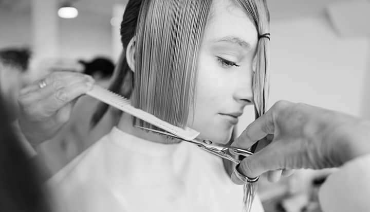 Strzyżenie prostych, mokrych włosów tuż pod brodą, wykonywane nożyczkami i grzebieniem przez stylistę. Ujęcie z profilu, w czerni i bieli.