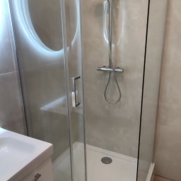 BM Construction - Nowoczesna kabina prysznicowa z przesuwnymi drzwiami, deszczownicą i baterią termostatyczną, obok umywalka z szafką w łazience wykończonej w jasnych odcieniach.