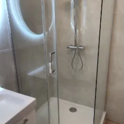 Nowoczesna kabina prysznicowa z przesuwnymi drzwiami, deszczownicą i baterią termostatyczną, obok umywalka z szafką w łazience wykończonej w jasnych odcieniach.