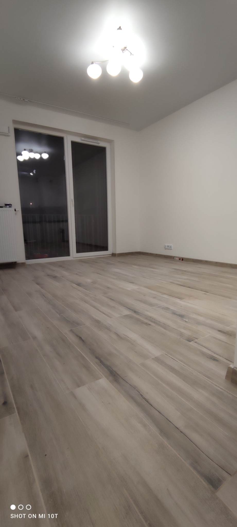 Pusty pokój z nową podłogą z paneli imitujących drewno, przesuwne drzwi balkonowe z białymi ramami, minimalistyczna lampa sufitowa z kulistymi kloszami, świeżo pomalowane ściany na biało.