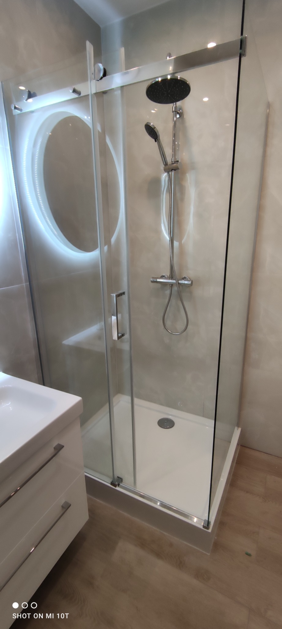 Nowoczesna kabina prysznicowa z przesuwnymi drzwiami, deszczownicą i baterią termostatyczną, obok umywalka z szafką w łazience wykończonej w jasnych odcieniach.