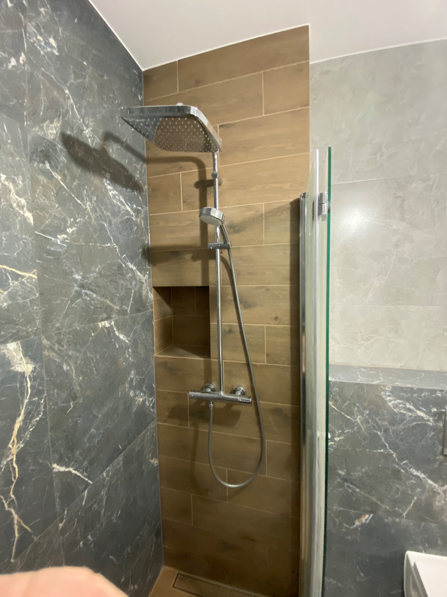 Kabina prysznicowa z deszczownicą i wnęką w brązowej ścianie z płytek imitujących drewno, obok ściana wyłożona marmurowymi płytkami w odcieniach szarości.