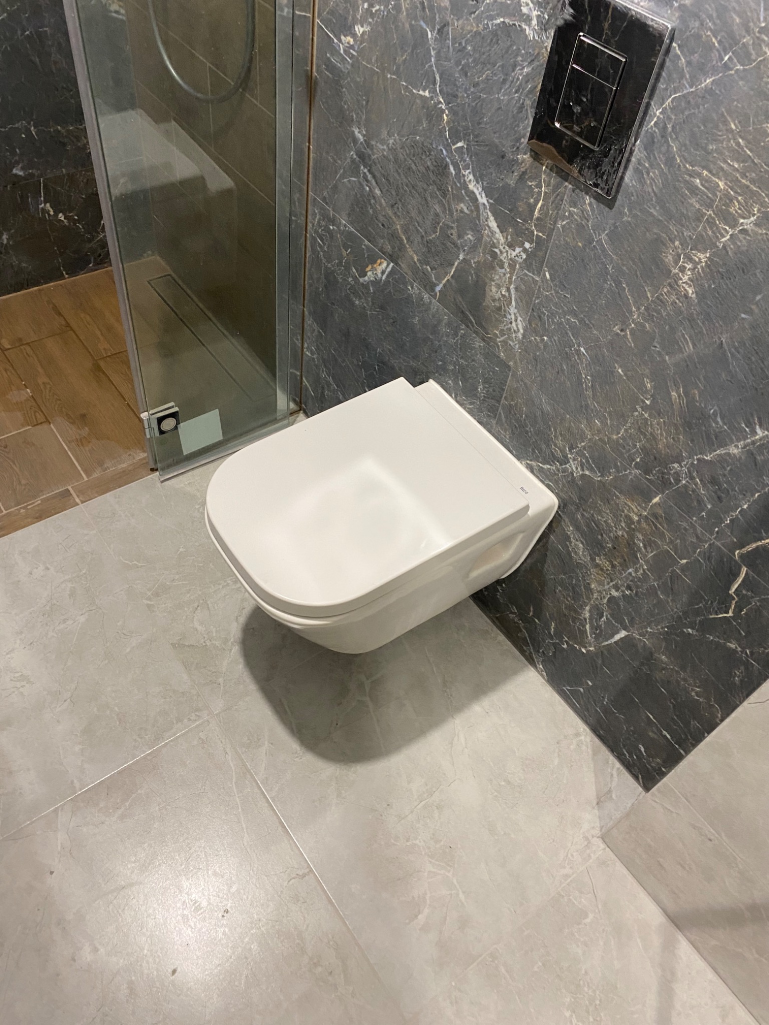 Nowoczesna toaleta podwieszana z czarnym marmurem na ścianie, szarą płytką podłogową i częściowo widoczną kabiną prysznicową z drewnianym akcentem pod nią.