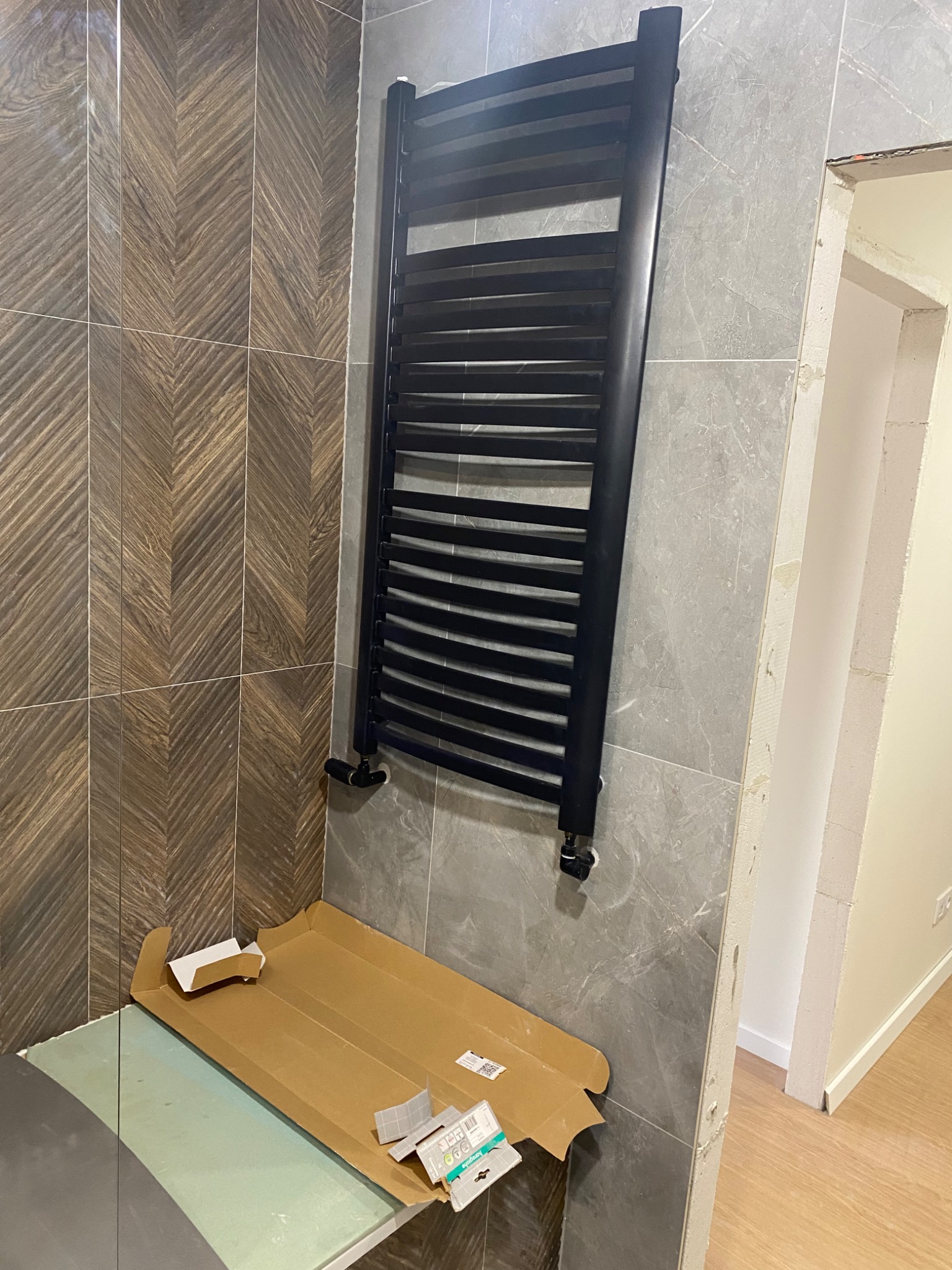 Czarny grzejnik łazienkowy drabinkowy zamontowany na szarej ścianie z płytek imitujących marmur, obok widoczna ściana z brązowych płytek ułożonych w jodełkę, na podłodze karton po opakowaniu...
