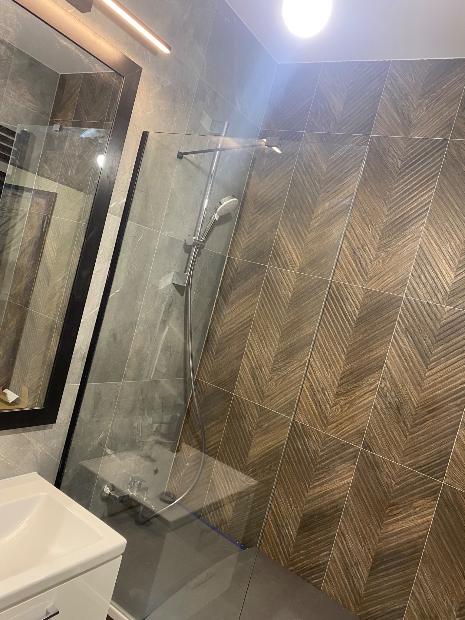 Nowoczesna kabina prysznicowa z szklaną ścianką, chromowaną armaturą i deszczownicą, wykończona płytkami imitującymi drewno ułożonymi w jodełkę na jednej ze ścian, oraz szarymi płytkami imitującymi...