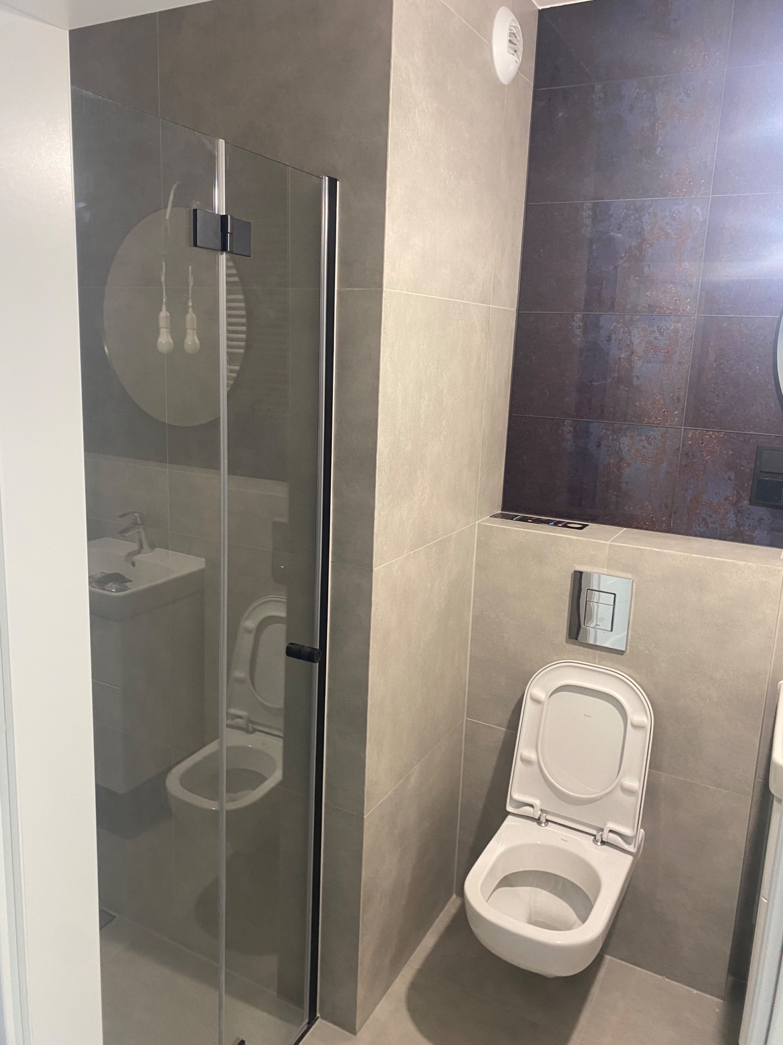 Nowoczesna łazienka z kabiną prysznicową ze szkła, wiszącą toaletą i umywalką, wykończona płytkami w odcieniach szarości i brązu.