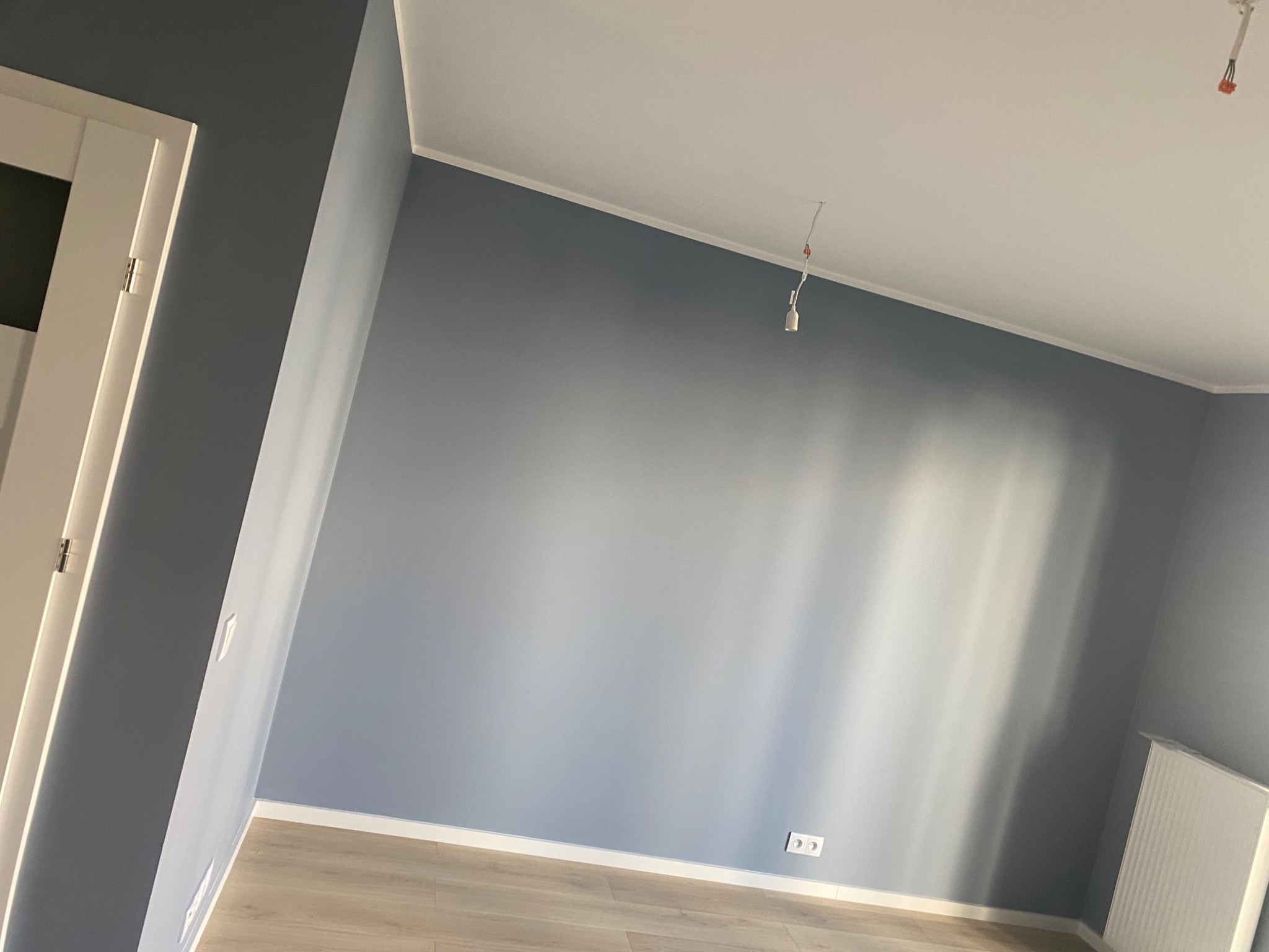 Świeżo pomalowane na szaro ściany w pustym pokoju z drewnianą podłogą i białymi listwami przypodłogowymi, widoczny biały grzejnik i wystający kabel od lampy.