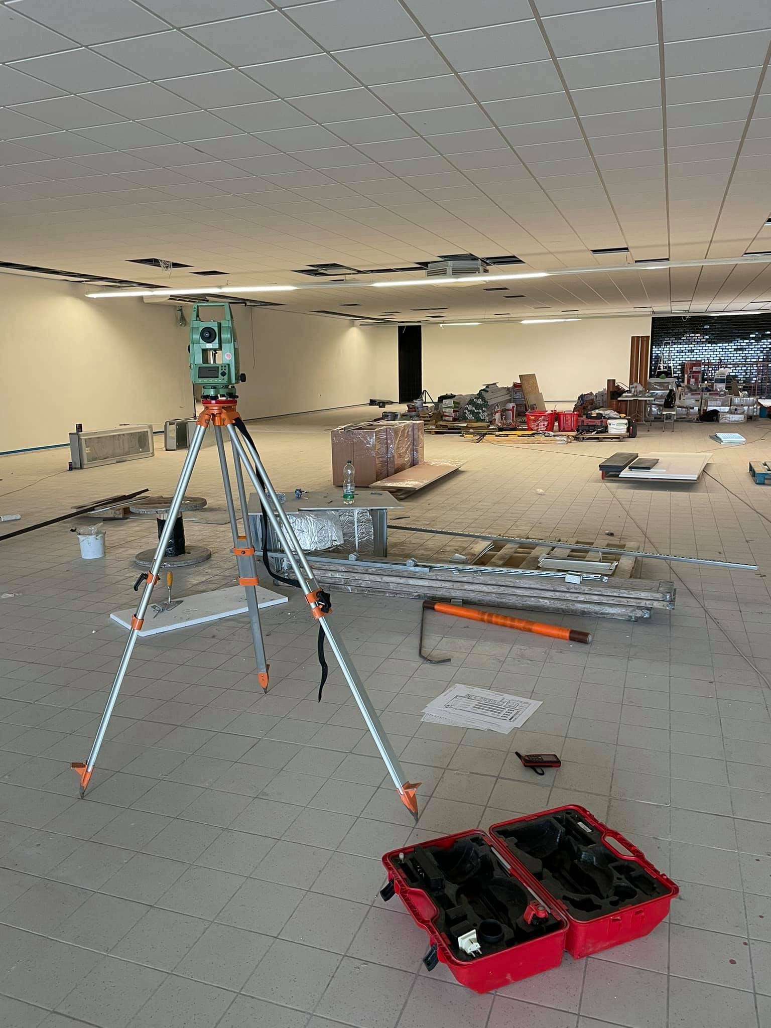 Zielony teodolit na statywie w pustym, remontowanym pomieszczeniu z kasetonowym sufitem, widoczna otwarta czerwona walizka na narzędzia i plany na podłodze.
