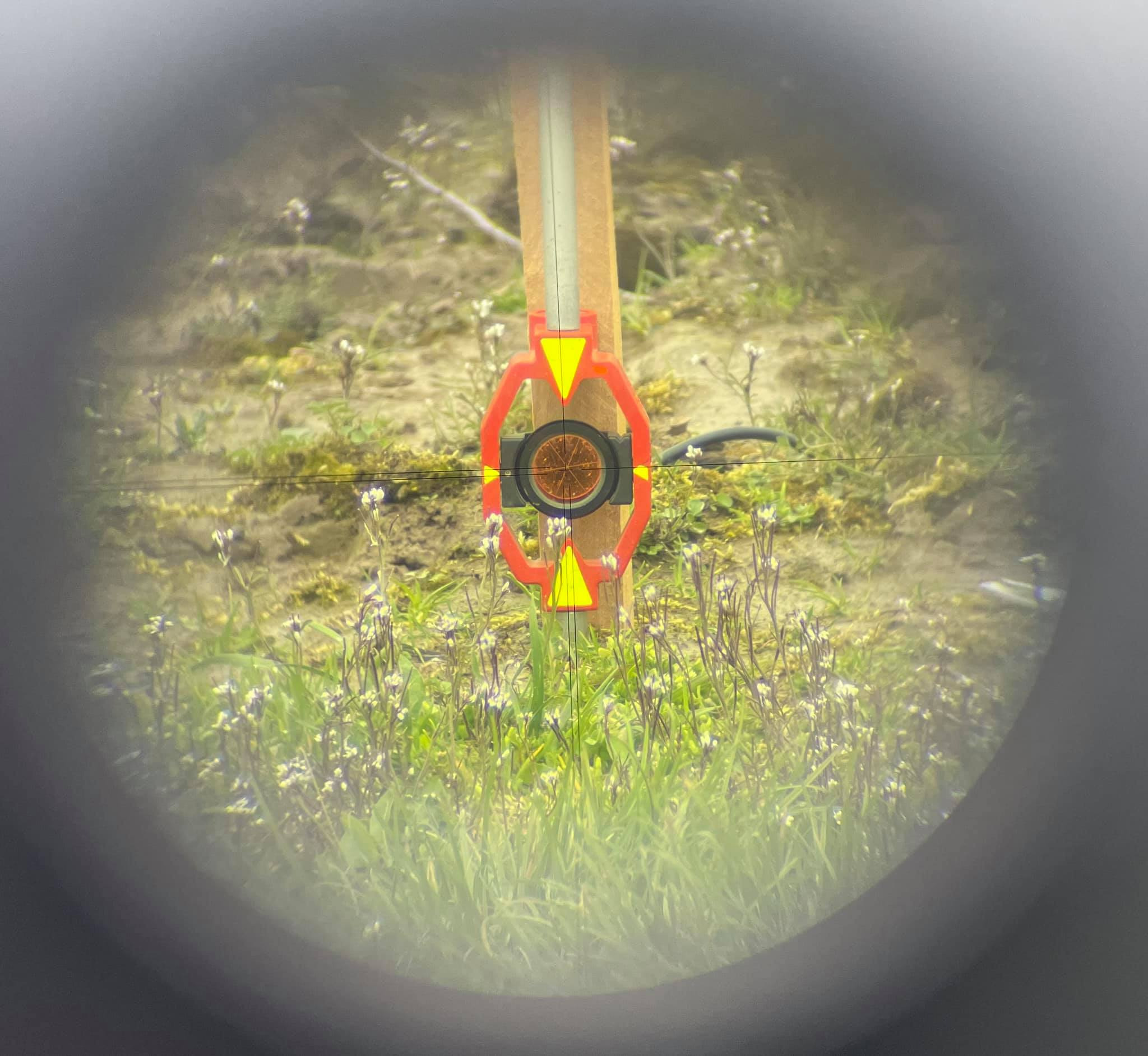 Widok przez niwelator na łatę geodezyjną z tarczą celowniczą na zarośniętym terenie.