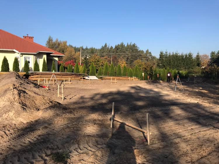 Prace geodezyjne na działce budowlanej: niwelator ustawiony na trójnogu, wytyczone punkty, widoczny dom z czerwonym dachem w tle.