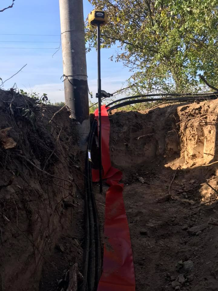 Prace ziemne: wykop z ułożonymi kablami zabezpieczonymi czerwoną folią, obok słup z zamontowanym odbiornikiem GPS na tyczce.