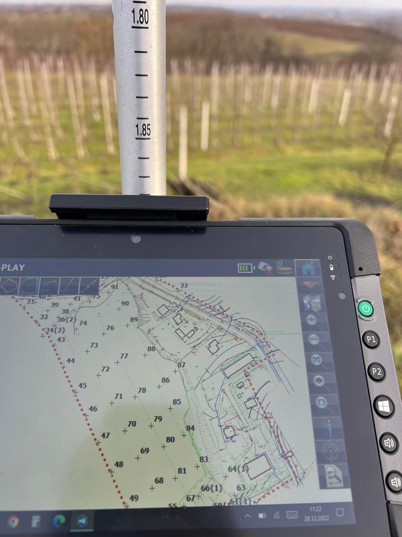 Ekran tabletu z uruchomionym oprogramowaniem do pomiarów geodezyjnych, w tle widoczny tyczką geodezyjną i winnica.