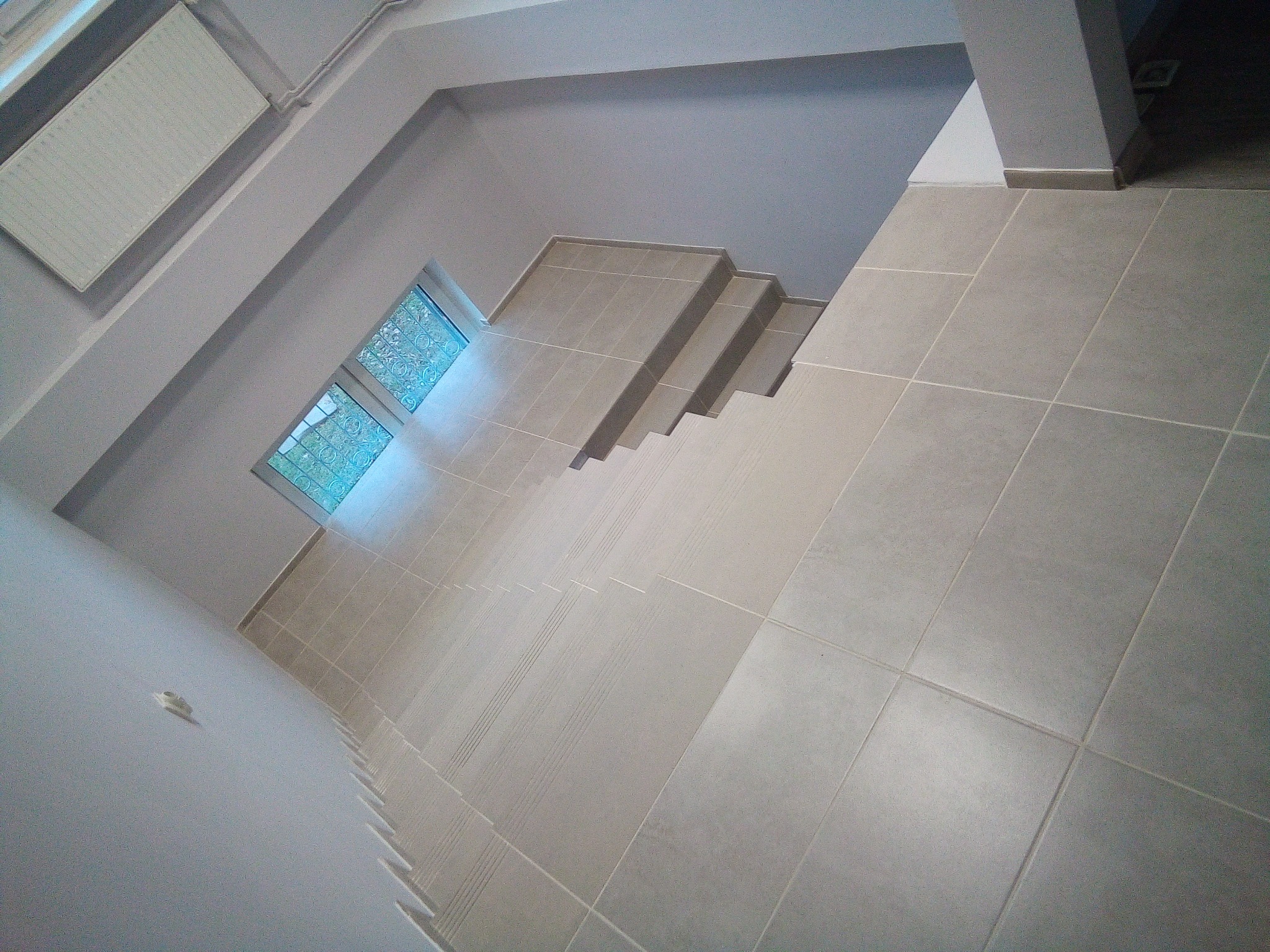 Klatka schodowa wyłożona jasnoszarymi płytkami ceramicznymi, widok z góry, z oknem po lewej stronie i grzejnikiem na ścianie.