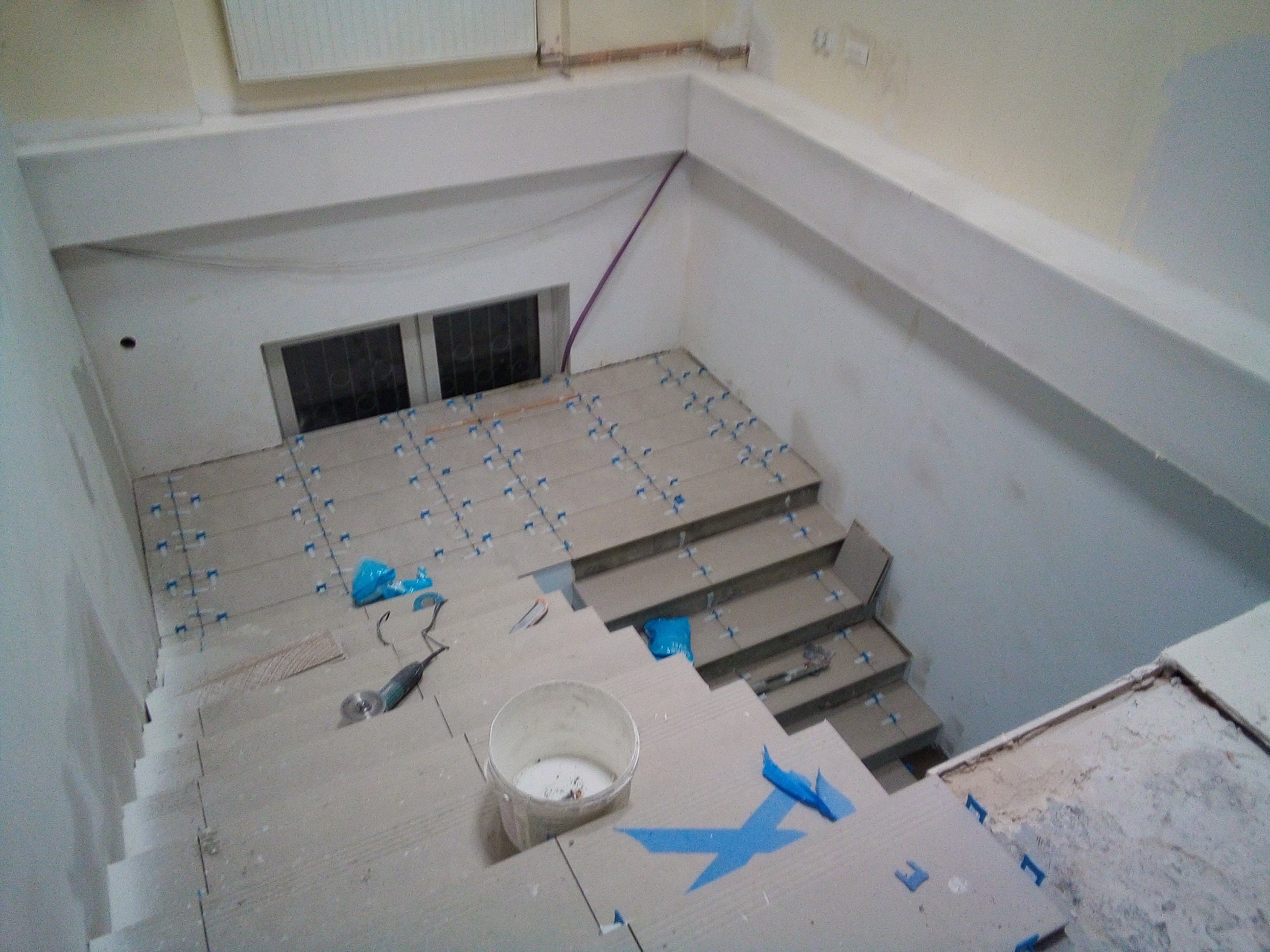 Schody w trakcie układania płytek, widoczne plastikowe krzyżyki dystansowe, wiadro z zaprawą i szlifierka kątowa, surowe ściany w tle.